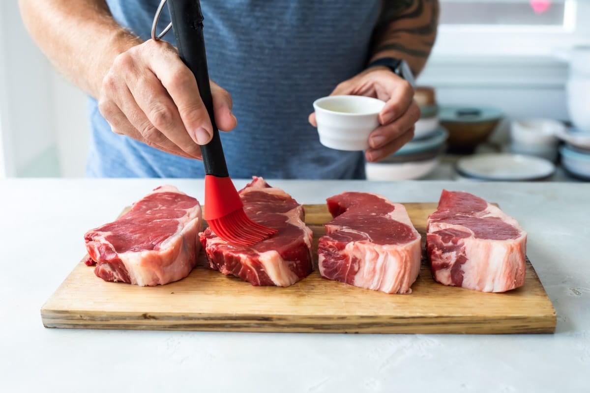 Brushing olive oil on raw New York strip steaks on a cutting board.