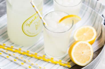 Lemonade in glasses on a silver platter with yellow and white straws and lemon garnish.