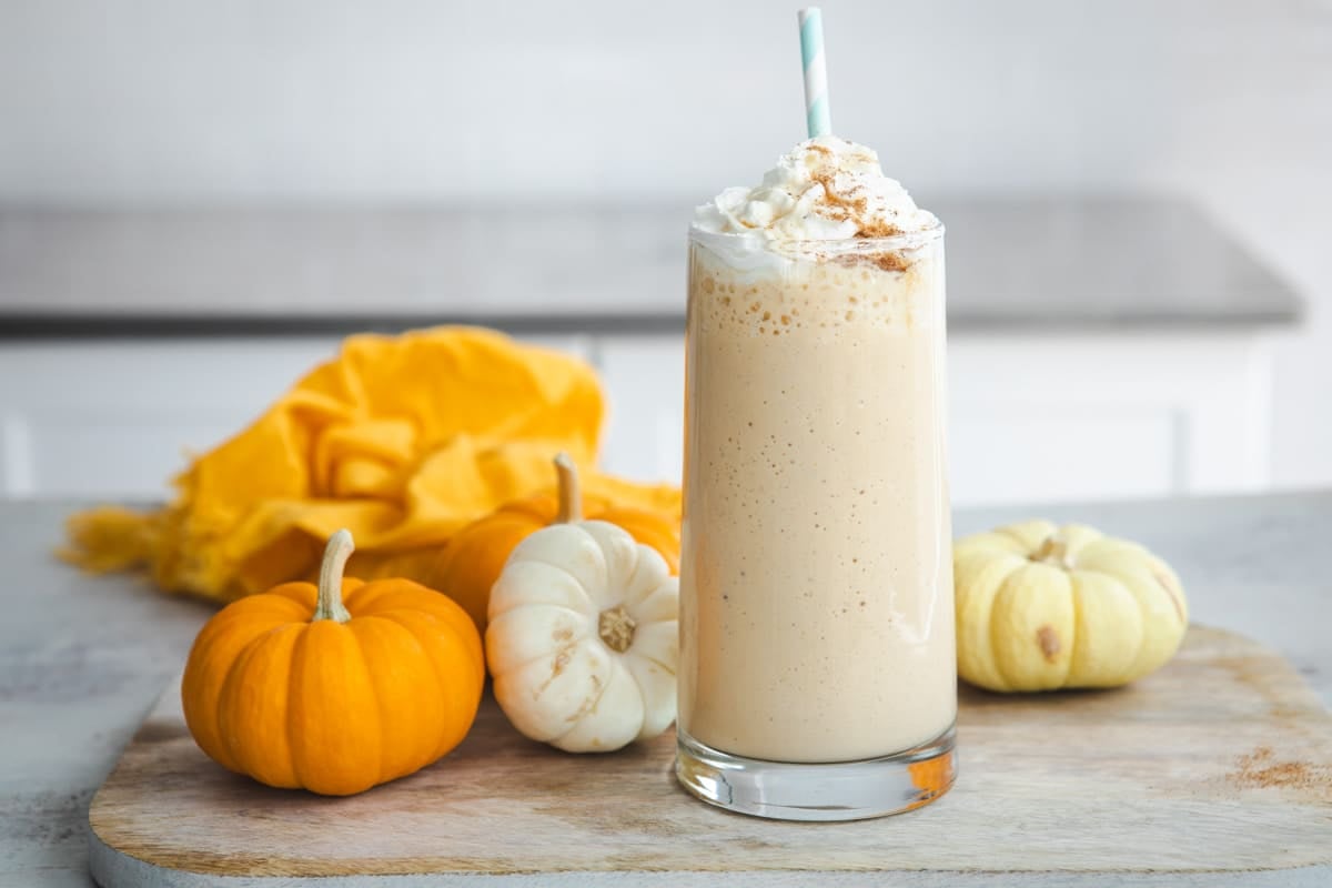 A pumpkin smoothie in a clear glass surrounded by small pumpkins.