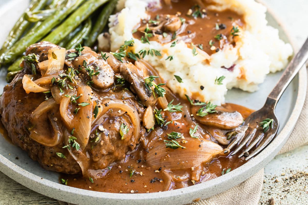 A salisbury steak on a plate with mashed potatoes, gravy, and green beans.